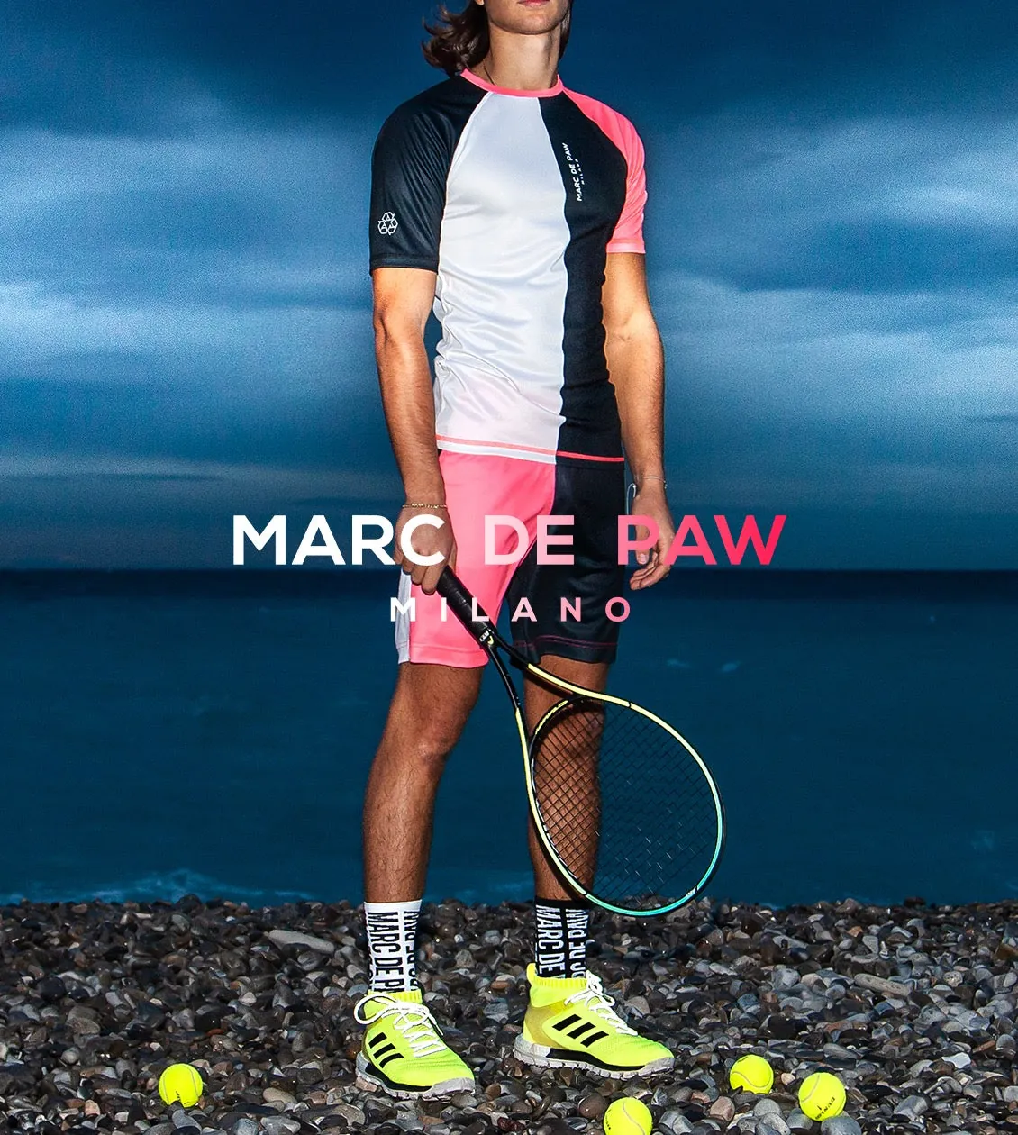 A male model wearing a white shirt and black shorts stands on a tennis court, holding a tennis racket in his hand. He is posing for a photo with several tennis balls around him, showcasing his passion for the sport with the Marc De Paw logo overlaid.