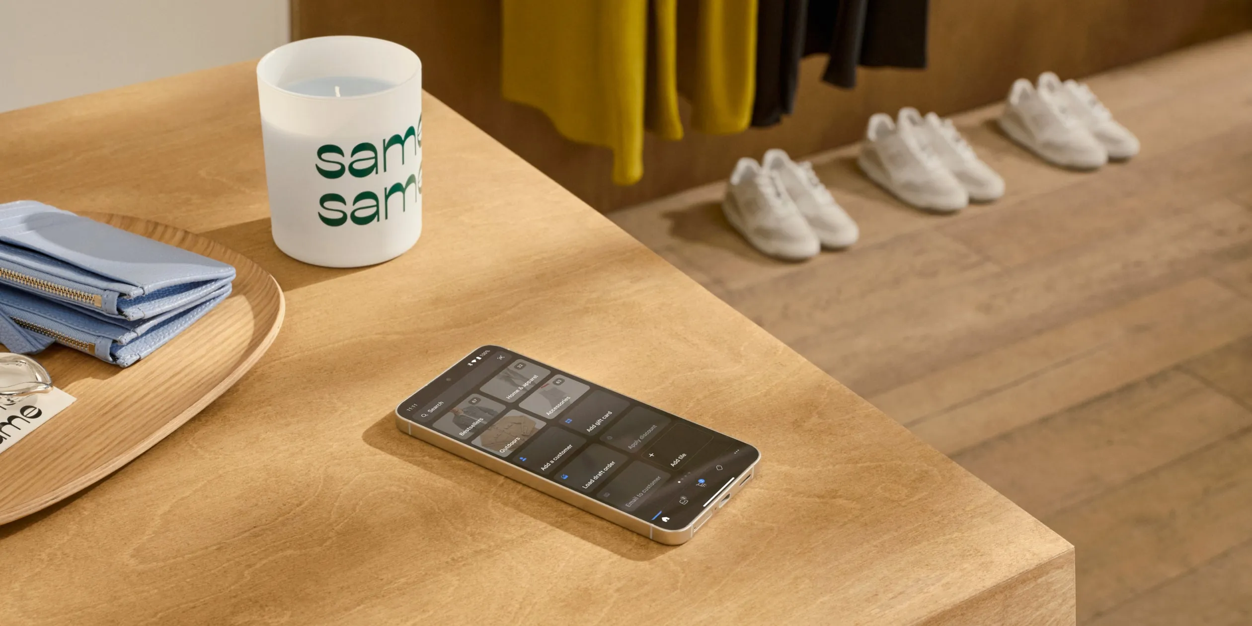 A close up of an Android device on a wood table with the PlatformDTC POS grid on its display. On the table is a candle with the store’s name, as well as other accessories. In the background, products including garments and shoes indicate a stylish retail store.