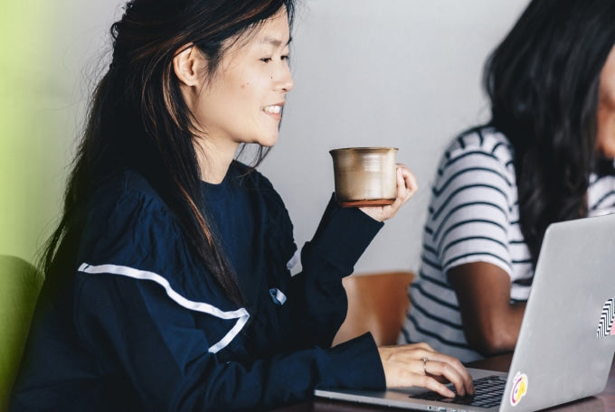 A woman sits in a cafe and smiles as she holds a coffee cup in one one hand, while she types on her laptop with her other.