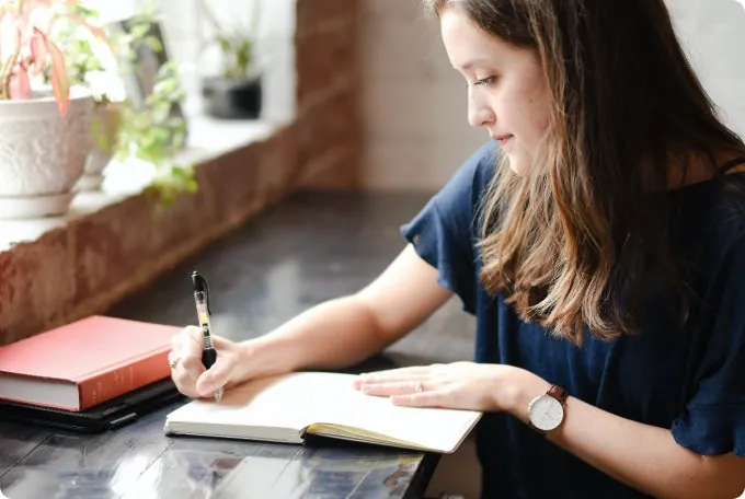 A woman sits at a well-lit desk and writes in her notebook.