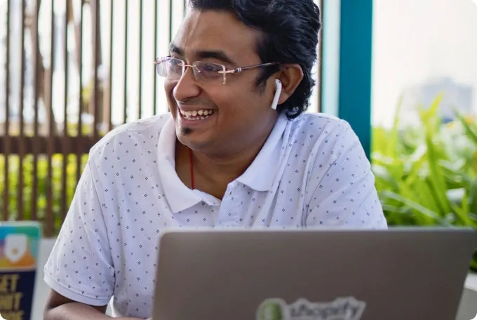 A man wearing wireless earbuds on, having a conversation while he sits facing his laptop.