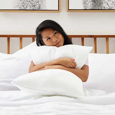 An image showing a woman sitting on a bed with white sheets, hugging a pillow.