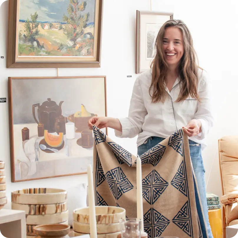 A woman wearing a white blouse and holding a patterned blanket, surrounded by paintings, candles and painted bowls