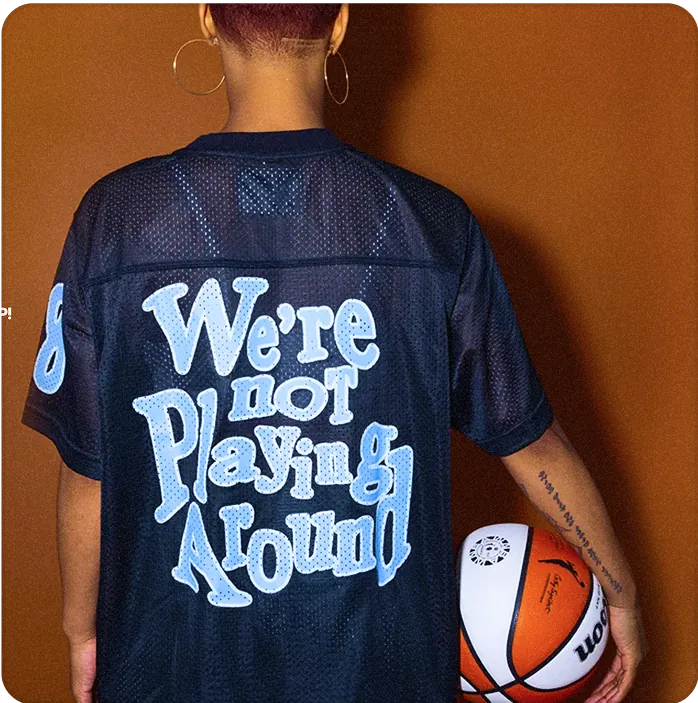 The back of a person in a navy t-shirt with baby blue writing holding a basketball.