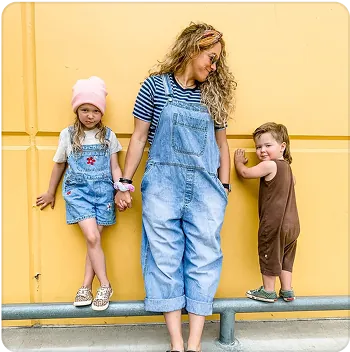 A woman in jean overalls standing with two children.